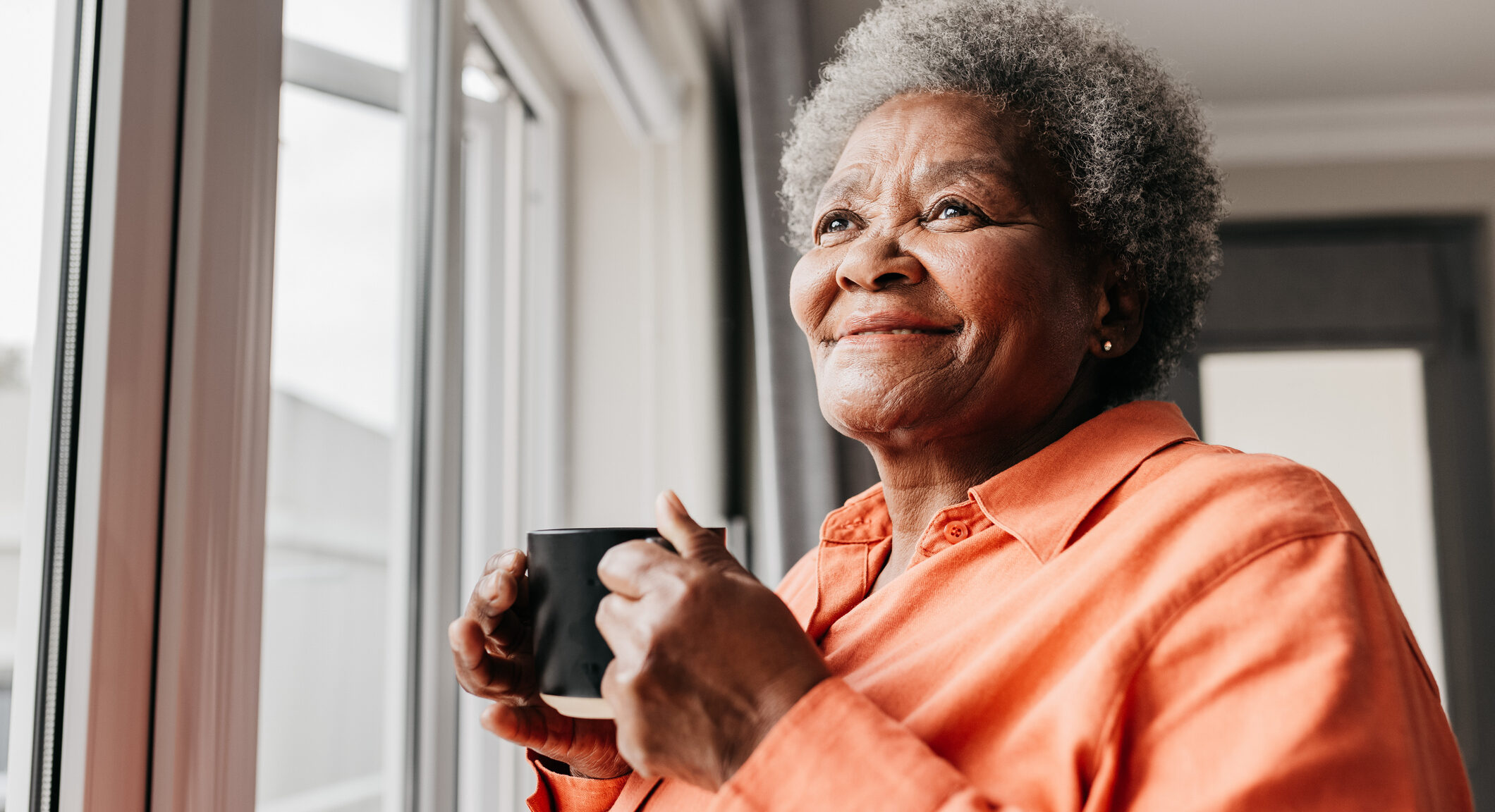 Senior woman holding mug, smiling and looking into distance beside window at home