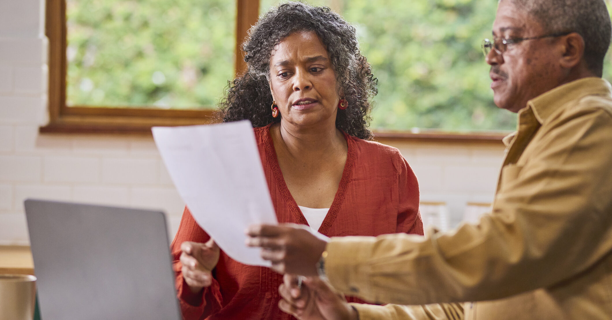 A concerned senior African American couple stands in their kitchen, seriously reviewing financial documents and looking at a laptop.
